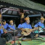 Traditional Indonesian music group performing on a covered outdoor stage at night, wearing blue shirts and orange headwraps.