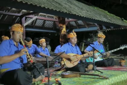 Traditional Indonesian music group performing on a covered outdoor stage at night, wearing blue shirts and orange headwraps.