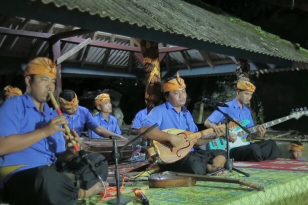 Traditional Indonesian music group performing on a covered outdoor stage at night, wearing blue shirts and orange headwraps.