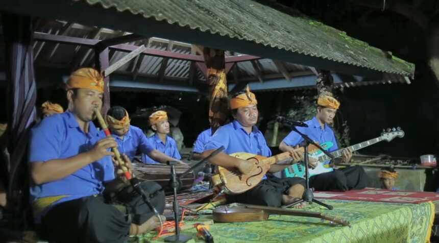 Traditional Indonesian music group performing on a covered outdoor stage at night, wearing blue shirts and orange headwraps.