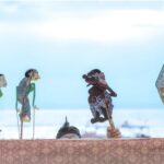 Colorful rod puppets on sticks stand above a patterned cloth, with a performer's eyes visible behind them, by a sea backdrop.