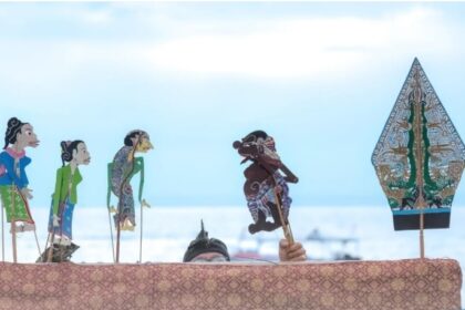 Colorful rod puppets on sticks stand above a patterned cloth, with a performer's eyes visible behind them, by a sea backdrop.