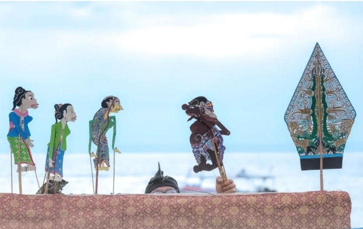 Colorful rod puppets on sticks stand above a patterned cloth, with a performer's eyes visible behind them, by a sea backdrop.