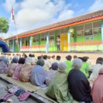 Group of students seated on mats outdoors, listening to a speaker on a small platform in front of a colorful school building.