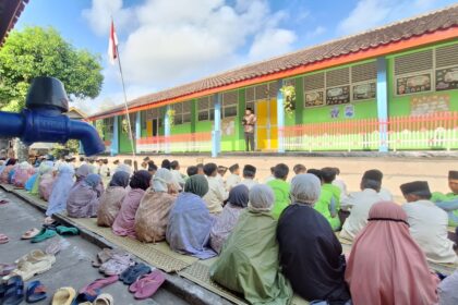 Group of students seated on mats outdoors, listening to a speaker on a small platform in front of a colorful school building.