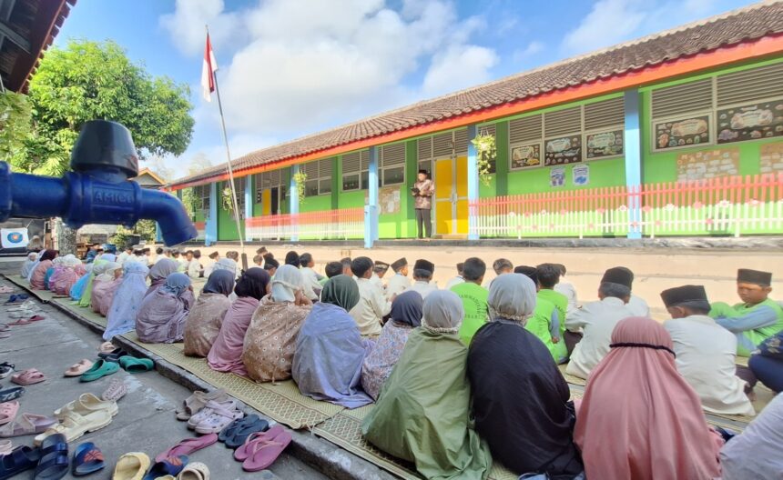 Group of students seated on mats outdoors, listening to a speaker on a small platform in front of a colorful school building.