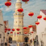 Sunlit street with a tall yellow minaret and red lanterns hanging above, white buildings on both sides, a scooter rider in the distance.