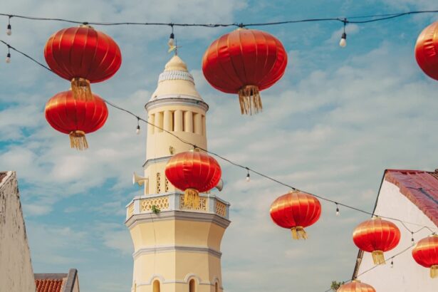 Sunlit street with a tall yellow minaret and red lanterns hanging above, white buildings on both sides, a scooter rider in the distance.