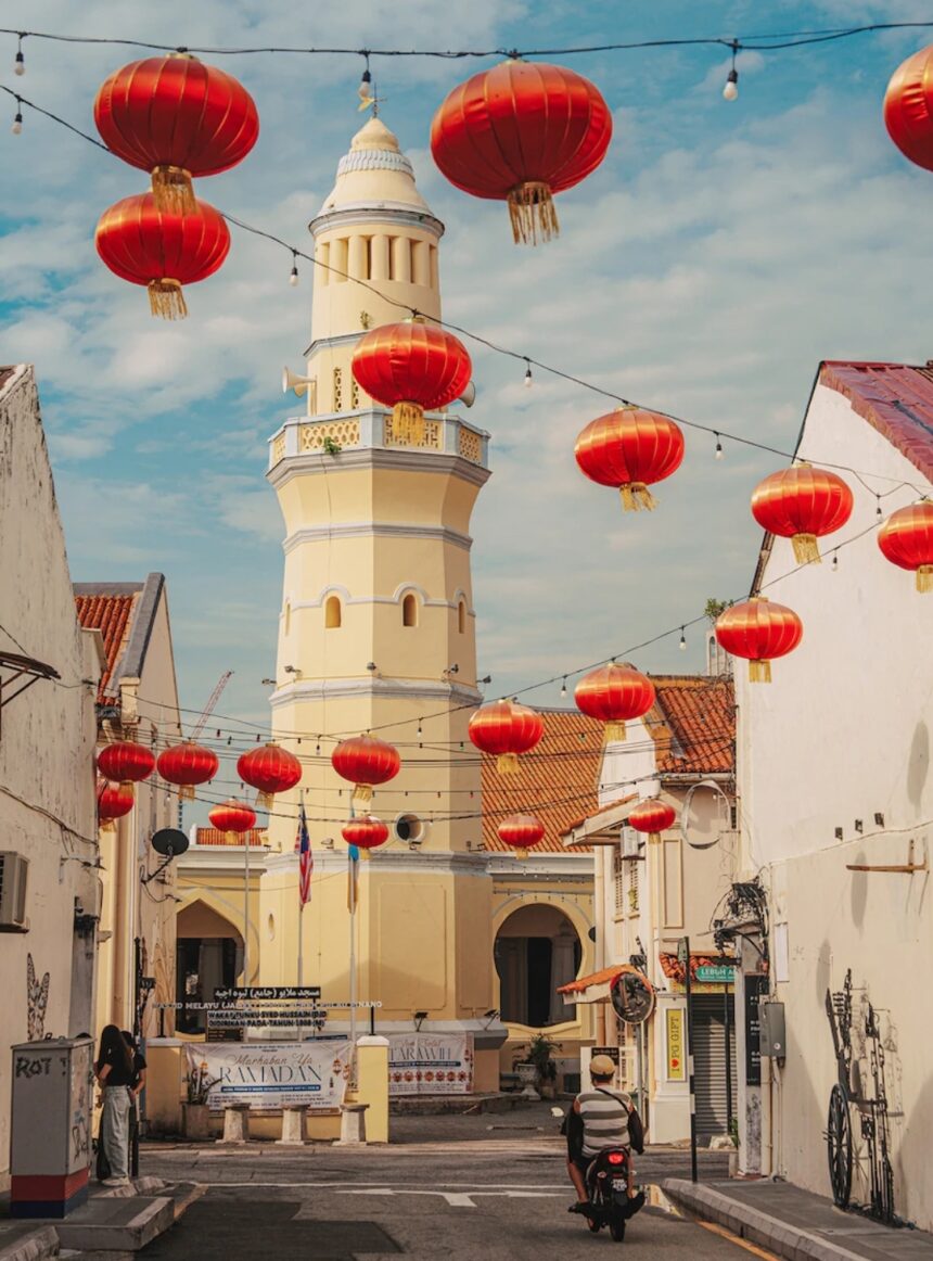 Sunlit street with a tall yellow minaret and red lanterns hanging above, white buildings on both sides, a scooter rider in the distance.