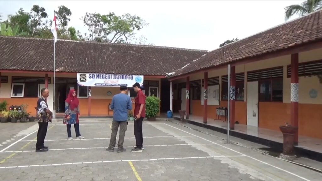 Screenshot_20261022_150313_Drive - Mabur.co Group of four adults conversing in a school courtyard; Indonesian flag on a tall pole and a banner reading 'SD Negeri Jatiroto' over an orange-brown building with a tiled roof.