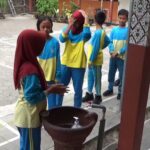 Group of schoolchildren in blue and yellow uniforms standing in a line outdoors near a water fountain, supervised by an adult in a hijab.