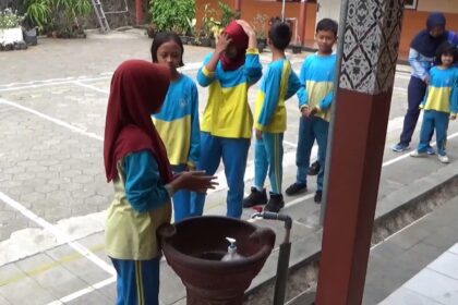 Group of schoolchildren in blue and yellow uniforms standing in a line outdoors near a water fountain, supervised by an adult in a hijab.
