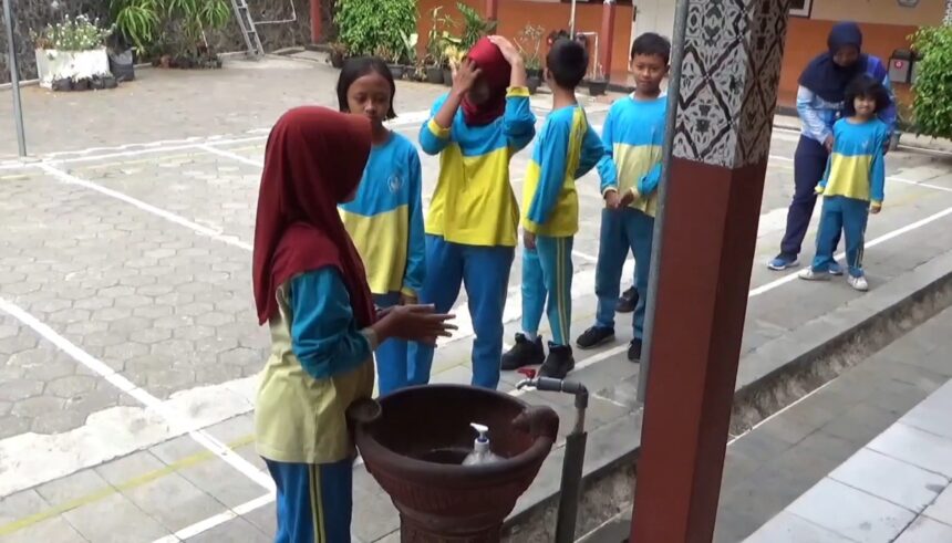 Group of schoolchildren in blue and yellow uniforms standing in a line outdoors near a water fountain, supervised by an adult in a hijab.