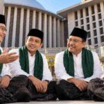 Four men in white shirts, black caps, and green sashes sit and chat outdoors in front of a large modern building.