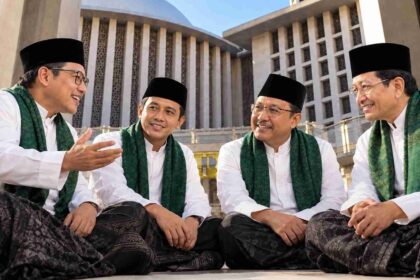Four men in white shirts, black caps, and green sashes sit and chat outdoors in front of a large modern building.