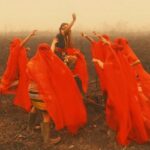 Group of people in bright red robes dancing in a smoke-filled, burnt field around a central figure raising an arm.