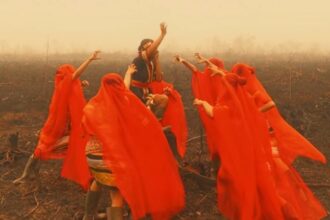 Group of people in bright red robes dancing in a smoke-filled, burnt field around a central figure raising an arm.