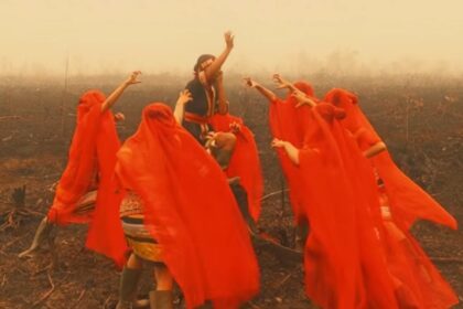 Group of people in bright red robes dancing in a smoke-filled, burnt field around a central figure raising an arm.