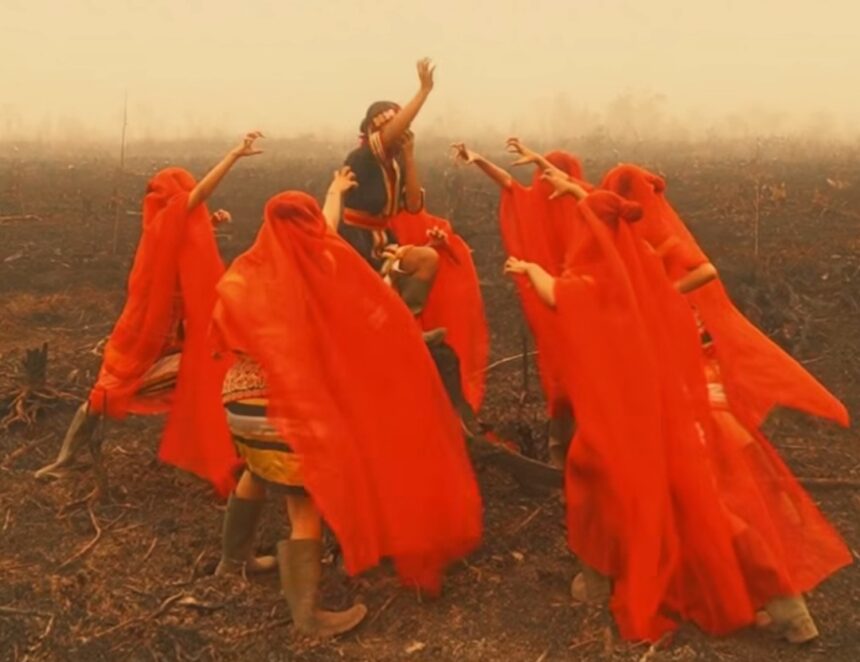 Group of people in bright red robes dancing in a smoke-filled, burnt field around a central figure raising an arm.