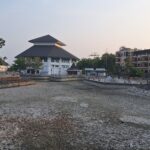 Riverside park with a tiered-roof white building, stone walls, and a boardwalk; people stroll along the water at dusk.