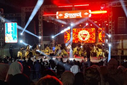 Stage at night with red lighting and a large screen reading 'Sitandi Fest'; dancers in gold and black costumes perform in front of a cheering crowd.