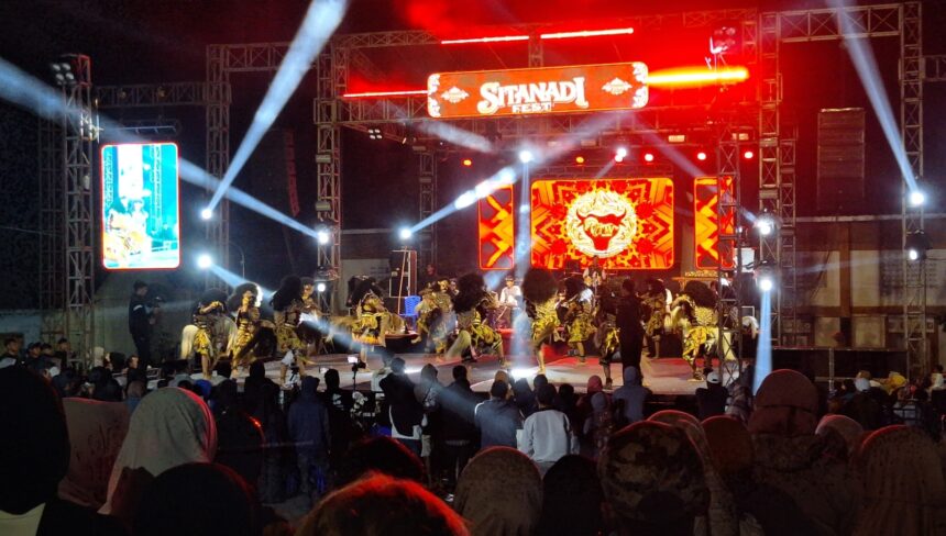Stage at night with red lighting and a large screen reading 'Sitandi Fest'; dancers in gold and black costumes perform in front of a cheering crowd.