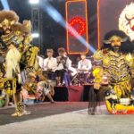 Group of performers in ornate gold Barong costumes on a stage, with spectators and a bright LED backdrop behind them.