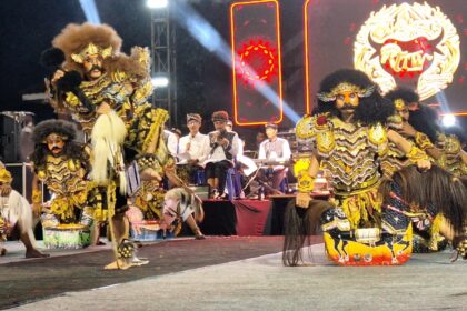Group of performers in ornate gold Barong costumes on a stage, with spectators and a bright LED backdrop behind them.