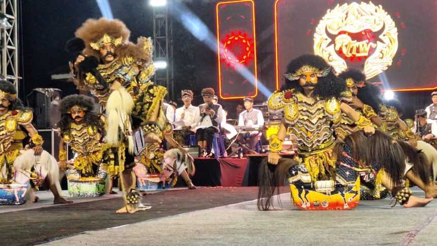 Group of performers in ornate gold Barong costumes on a stage, with spectators and a bright LED backdrop behind them.