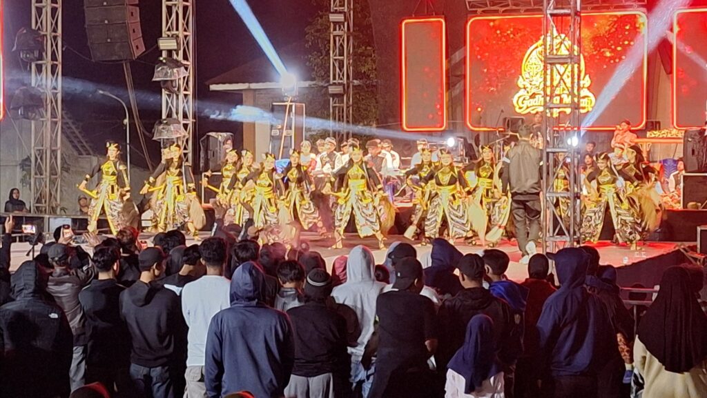 Group of dancers in gold and black costumes performing on a lit stage with a cheering crowd watching at night.