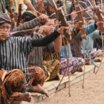 Group kneeling on mats, drawing wooden bows in an outdoor archery practice run.