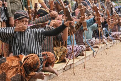 Group kneeling on mats, drawing wooden bows in an outdoor archery practice run.
