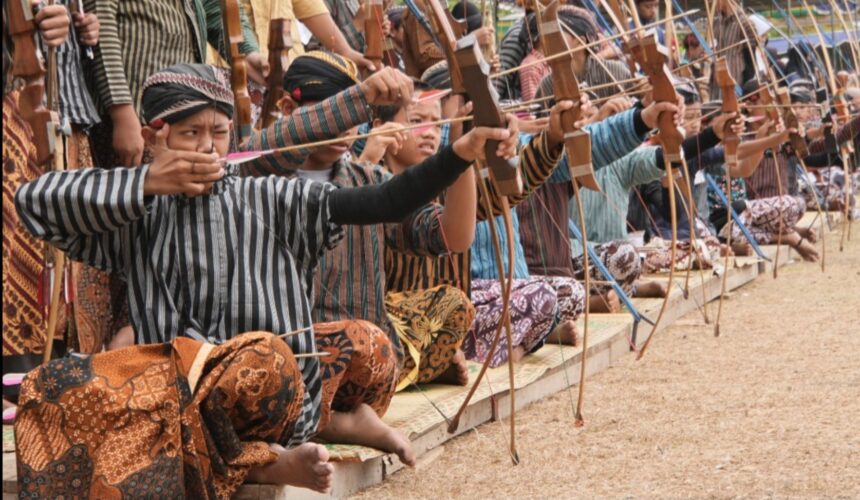 Group kneeling on mats, drawing wooden bows in an outdoor archery practice run.