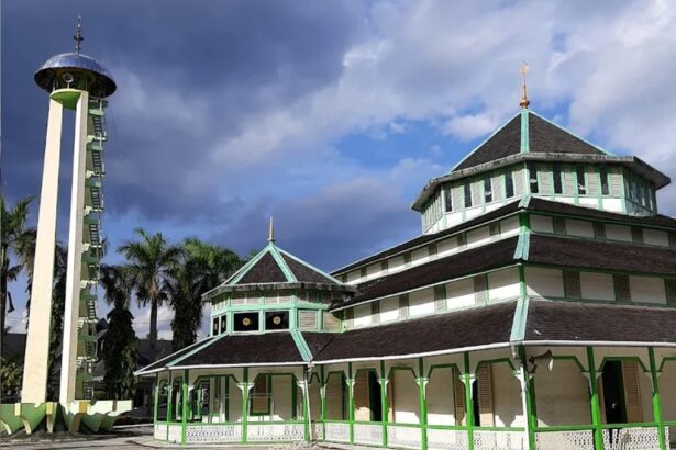 Green and white multi-tiered temple with a tall vertical tower on the left, palm trees in the background under a cloudy sky.