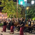Nighttime street performance: four women in black tops and red patterned skirts dance while a small musical ensemble plays traditional instruments nearby.