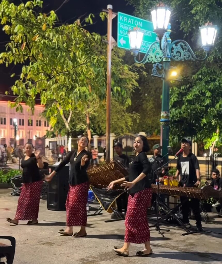 Nighttime street performance: four women in black tops and red patterned skirts dance while a small musical ensemble plays traditional instruments nearby.