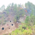 Hilly rock face with square cave openings and a wildfire burning among trees on the slope.