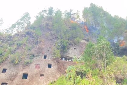 Hilly rock face with square cave openings and a wildfire burning among trees on the slope.
