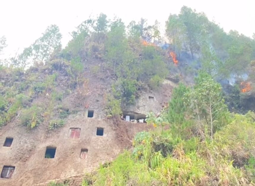 Hilly rock face with square cave openings and a wildfire burning among trees on the slope.