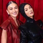 Two women smile and pose together in front of a red curtain; one wears a red leather jacket, the other a black sequined hooded dress.