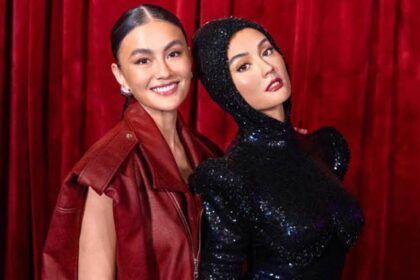 Two women smile and pose together in front of a red curtain; one wears a red leather jacket, the other a black sequined hooded dress.
