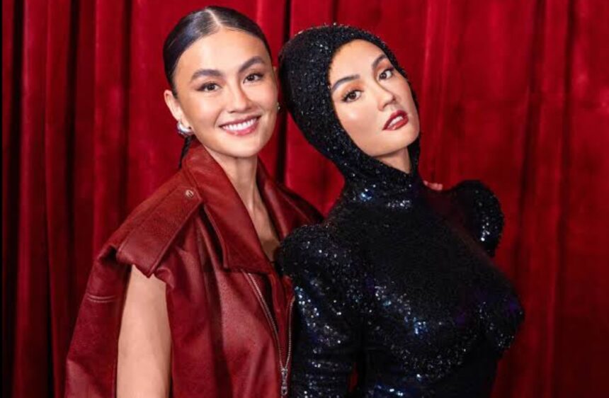 Two women smile and pose together in front of a red curtain; one wears a red leather jacket, the other a black sequined hooded dress.