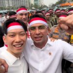 Two men in white shirts and red-white headbands selfie with a crowd behind them at an outdoor event, police visible in the background.