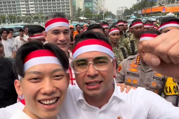 Two men in white shirts and red-white headbands selfie with a crowd behind them at an outdoor event, police visible in the background.