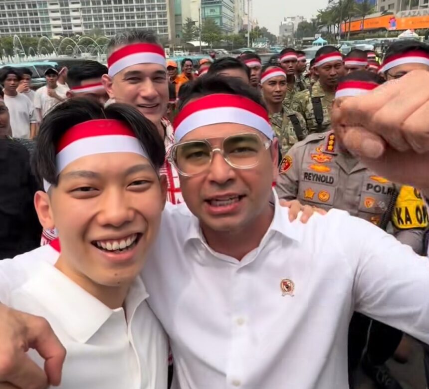 Two men in white shirts and red-white headbands selfie with a crowd behind them at an outdoor event, police visible in the background.