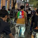 Group of people riding bikes down a lit urban street at night; the central rider wears a tan shirt with a rainbow patch showing a white raised-hand and text on the back.
