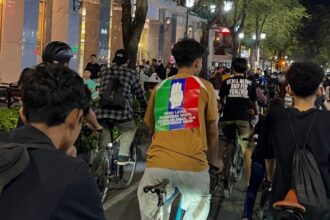 Group of people riding bikes down a lit urban street at night; the central rider wears a tan shirt with a rainbow patch showing a white raised-hand and text on the back.