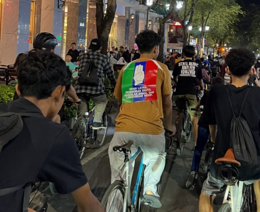 Group of people riding bikes down a lit urban street at night; the central rider wears a tan shirt with a rainbow patch showing a white raised-hand and text on the back.