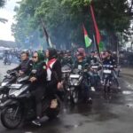 Group of riders on motorcycles in a street protest, several carrying Palestinian flags amid light smoke and trees in the background.