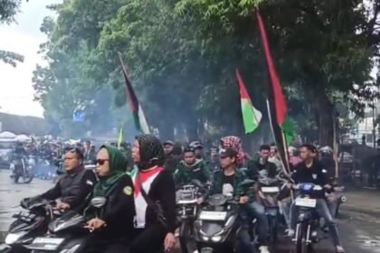 Group of riders on motorcycles in a street protest, several carrying Palestinian flags amid light smoke and trees in the background.